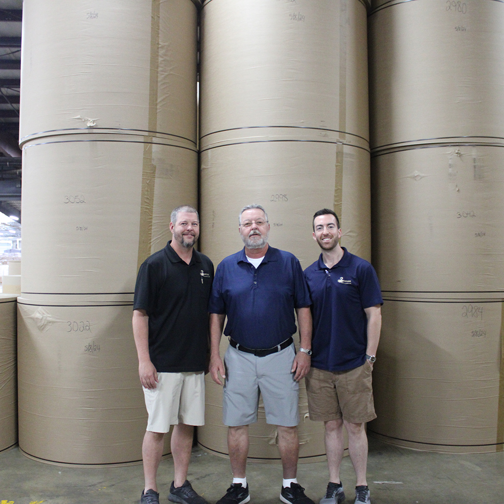 Three men standing in front of rolled up corrugated paper