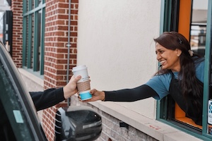 A cheerful BIGGBY COFFEE barista handing a drink to a customer at a brick drive-thru window.