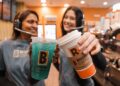 Two BIGGBY COFFEE baristas smiling and holding up a teal-blue iced drink and a hot specialty coffee in a branded orange-and-white cup