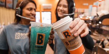 Two BIGGBY COFFEE baristas smiling and holding up a teal-blue iced drink and a hot specialty coffee in a branded orange-and-white cup