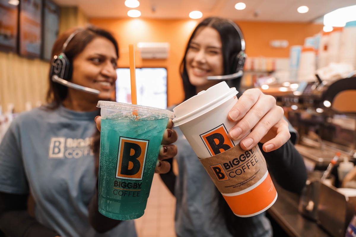 Two BIGGBY COFFEE baristas smiling and holding up a teal-blue iced drink and a hot specialty coffee in a branded orange-and-white cup