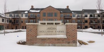 Holliday Farms Senior Living sign in front of a three-story brick apartment building, with snow covering the ground and overcast winter skies.