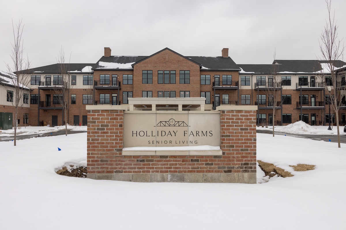 Holliday Farms Senior Living sign in front of a three-story brick apartment building, with snow covering the ground and overcast winter skies.