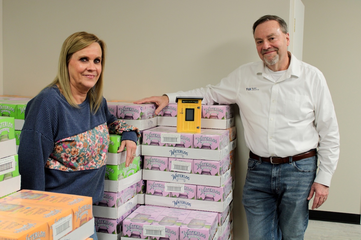 Founders of the Feeding Team nonprofit pose with stacks of donated beverages and a model of their "no-questions-asked" yellow food pantry.