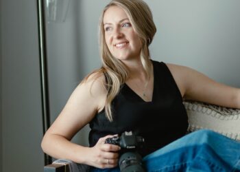 Portrait of a smiling woman in a black tank top and jeans holding a Canon camera while seated in a cozy armchair
