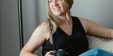 Portrait of a smiling woman in a black tank top and jeans holding a Canon camera while seated in a cozy armchair