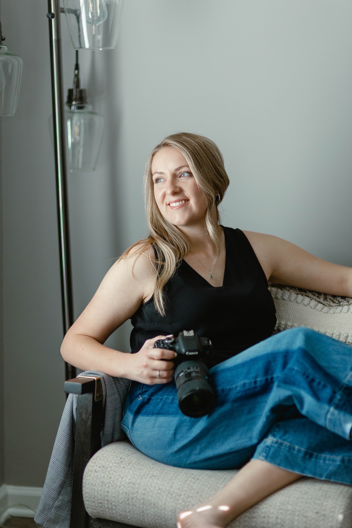 Portrait of a smiling woman in a black tank top and jeans holding a Canon camera while seated in a cozy armchair