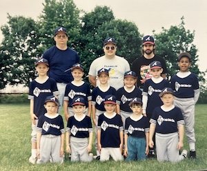 A group photo of a youth baseball team and three coaches posing on a grassy field in front of a line of trees