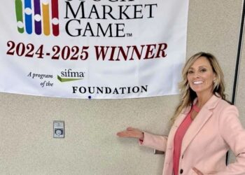 A woman with blonde hair, wearing a light pink blazer over a coral pink top, stands in front of a neutral-colored wall. She is smiling and gesturing with one hand toward a white banner that reads "THE STOCK MARKET GAME 2024-2025 WINNER." The banner also features a colorful bar graph logo and the SIFMA Foundation logo.