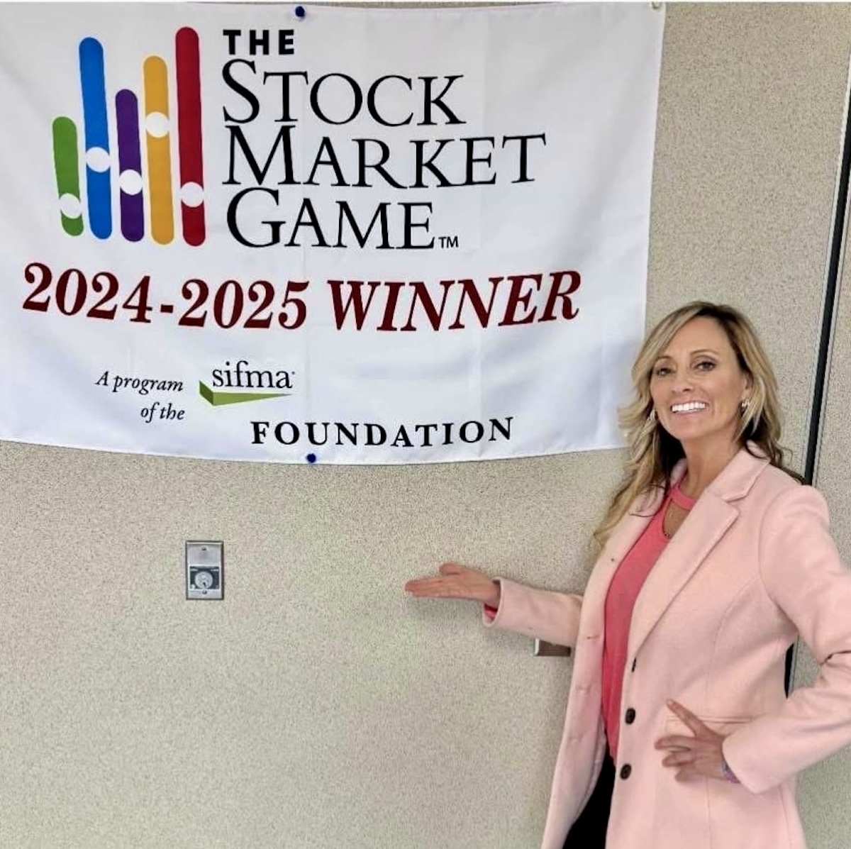 A woman with blonde hair, wearing a light pink blazer over a coral pink top, stands in front of a neutral-colored wall. She is smiling and gesturing with one hand toward a white banner that reads "THE STOCK MARKET GAME 2024-2025 WINNER." The banner also features a colorful bar graph logo and the SIFMA Foundation logo.