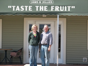 Two people, a man and a woman, stand smiling in front of the white door of a store.The woman, on the left, is wearing a dark green hooded sweatshirt and blue jeans. The man, on the right, wears a light blue button-down shirt tucked into blue jeans. Above them, a green awning features the white text "TASTE THE FRUIT" with a smaller "JAMS & JELLIES" logo above it. To the left, a small black bistro table and chair are visible on the patio.
