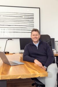 A smiling man in a navy blue quarter-zip sweater and light grey trousers sits at a large, live-edge wooden desk in a modern office. He leans comfortably against the desk, where a laptop is open. Behind him, a sleek black desk lamp and dual computer monitors are visible. On the wall hangs a large, minimalist piece of art featuring horizontal black lines on a white background.
