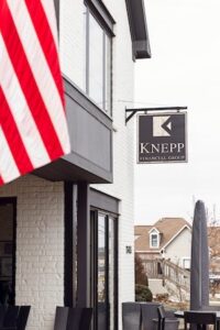 The exterior of a modern, two-story white brick building features the hanging sign for "KNEPP FINANCIAL GROUP." The black, square sign displays a white geometric logo above the company name in all-capital letters. To the left, a portion of a large American flag with its red and white stripes is visible in the foreground. In the background, a residential house with a brown roof and a wooden deck can be seen under a bright, overcast sky.