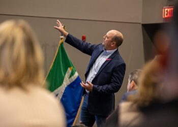 A candid, over-the-shoulder shot of Fishers Mayor Scott Fadness speaking at an indoor event. He is wearing a navy blue blazer, a light blue button-down shirt, and jeans. He is gesturing with his right hand toward the side, while his left hand is open near his waist as he addresses the audience. In the background, a green, white, and blue flag with a stylized "X" pattern—the official city flag of Fishers, Indiana—is visible. An illuminated red "EXIT" sign is mounted on the wall to the right. The foreground shows the blurred backs of several audience members' heads.