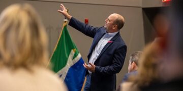 A candid, over-the-shoulder shot of Fishers Mayor Scott Fadness speaking at an indoor event. He is wearing a navy blue blazer, a light blue button-down shirt, and jeans. He is gesturing with his right hand toward the side, while his left hand is open near his waist as he addresses the audience. In the background, a green, white, and blue flag with a stylized "X" pattern—the official city flag of Fishers, Indiana—is visible. An illuminated red "EXIT" sign is mounted on the wall to the right. The foreground shows the blurred backs of several audience members' heads.