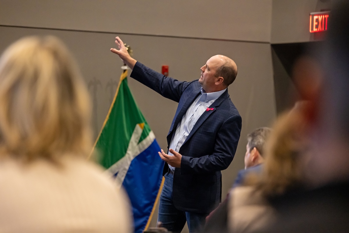 A candid, over-the-shoulder shot of Fishers Mayor Scott Fadness speaking at an indoor event. He is wearing a navy blue blazer, a light blue button-down shirt, and jeans. He is gesturing with his right hand toward the side, while his left hand is open near his waist as he addresses the audience. In the background, a green, white, and blue flag with a stylized "X" pattern—the official city flag of Fishers, Indiana—is visible. An illuminated red "EXIT" sign is mounted on the wall to the right. The foreground shows the blurred backs of several audience members' heads.