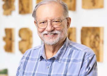A medium, waist-up portrait of an older man with gray hair, a short gray beard, and glasses, smiling warmly at the camera. He is wearing a light blue and white plaid button-down shirt. He stands in a brightly lit room with a clean, modern aesthetic. In the background, several rectangular pieces of natural wood art with dark knots and grain patterns are mounted on a white wall in a grid-like arrangement. A dark gray sofa with a yellow cushion is partially visible to the left.