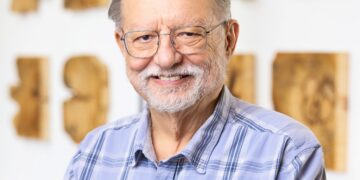 A medium, waist-up portrait of an older man with gray hair, a short gray beard, and glasses, smiling warmly at the camera. He is wearing a light blue and white plaid button-down shirt. He stands in a brightly lit room with a clean, modern aesthetic. In the background, several rectangular pieces of natural wood art with dark knots and grain patterns are mounted on a white wall in a grid-like arrangement. A dark gray sofa with a yellow cushion is partially visible to the left.