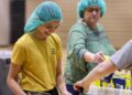A smiling young girl with dark hair, wearing a teal hairnet and a yellow "HAPPY CAMPER" t-shirt, works at a food packing station in a brightly lit gymnasium. She is focused on placing items into a plastic container. In the background, an older woman in a green patterned top and a matching teal hairnet reaches for a yellow food package. A third person's arm is visible on the right, also assisting with the packing process. Several yellow bags of food are stacked on a table in the foreground.