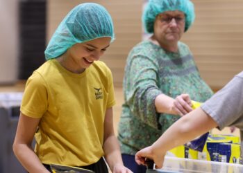 A smiling young girl with dark hair, wearing a teal hairnet and a yellow "HAPPY CAMPER" t-shirt, works at a food packing station in a brightly lit gymnasium. She is focused on placing items into a plastic container. In the background, an older woman in a green patterned top and a matching teal hairnet reaches for a yellow food package. A third person's arm is visible on the right, also assisting with the packing process. Several yellow bags of food are stacked on a table in the foreground.