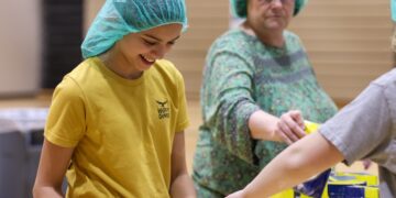 A smiling young girl with dark hair, wearing a teal hairnet and a yellow "HAPPY CAMPER" t-shirt, works at a food packing station in a brightly lit gymnasium. She is focused on placing items into a plastic container. In the background, an older woman in a green patterned top and a matching teal hairnet reaches for a yellow food package. A third person's arm is visible on the right, also assisting with the packing process. Several yellow bags of food are stacked on a table in the foreground.