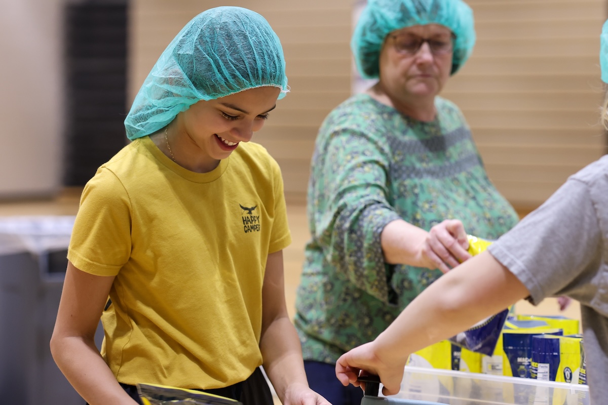 A smiling young girl with dark hair, wearing a teal hairnet and a yellow "HAPPY CAMPER" t-shirt, works at a food packing station in a brightly lit gymnasium. She is focused on placing items into a plastic container. In the background, an older woman in a green patterned top and a matching teal hairnet reaches for a yellow food package. A third person's arm is visible on the right, also assisting with the packing process. Several yellow bags of food are stacked on a table in the foreground.