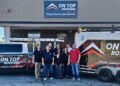 A team of eight employees from On Top Roofing stands together in front of their office building, flanked by a branded white pickup truck and a matching trailer.