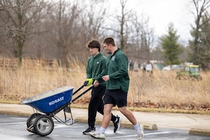 Two young men, likely students from Westfield High School given their matching dark green shirts featuring a white "W" logo, work together to push a blue Kobalt wheelbarrow across a paved area. The student on the left, with dark wavy hair, is dressed in black athletic pants and green gloves, while the student on the right, in black shorts and dark sneakers, smiles as he walks alongside. In the background, a field of tall, dry grass and several leafless trees under an overcast sky suggest a late winter or early spring day. A small green tractor is partially visible in the distance.