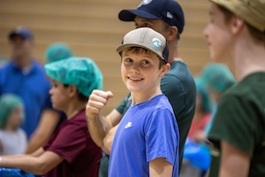 A smiling young boy in a bright blue t-shirt and a gray and black trucker hat gives a thumbs-up to the camera. He is standing in a large gymnasium during a community service event. In the background, other children and adults are visible, some wearing teal hairnets and participating in a meal-packing activity. The boy's cheerful expression and gesture highlight the positive atmosphere of the volunteer effort.