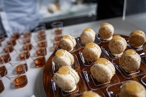 A close-up, high-angle shot of a catering display featuring rows of round, cream-colored pastries topped with a golden gel or jam. The pastries are individually served in small, clear square dishes on a wooden tray. To the left, numerous small clear cups filled with an amber-colored liquid are neatly arranged on a white tabletop. The background is softly blurred, showing people in a brightly lit, modern setting.