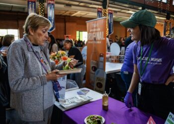 A woman in a grey fleece jacket samples a small salad from a cardboard tray at an indoor food event. She stands across a purple-clothed table from a representative in a green cap and purple "80 Acres Farms" t-shirt, who is smiling and talking to her. In the background, a tall orange banner for "Tito's Handmade Vodka" is visible among a crowd of people in a large, industrial-style hall.