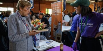 A woman in a grey fleece jacket samples a small salad from a cardboard tray at an indoor food event. She stands across a purple-clothed table from a representative in a green cap and purple "80 Acres Farms" t-shirt, who is smiling and talking to her. In the background, a tall orange banner for "Tito's Handmade Vodka" is visible among a crowd of people in a large, industrial-style hall.