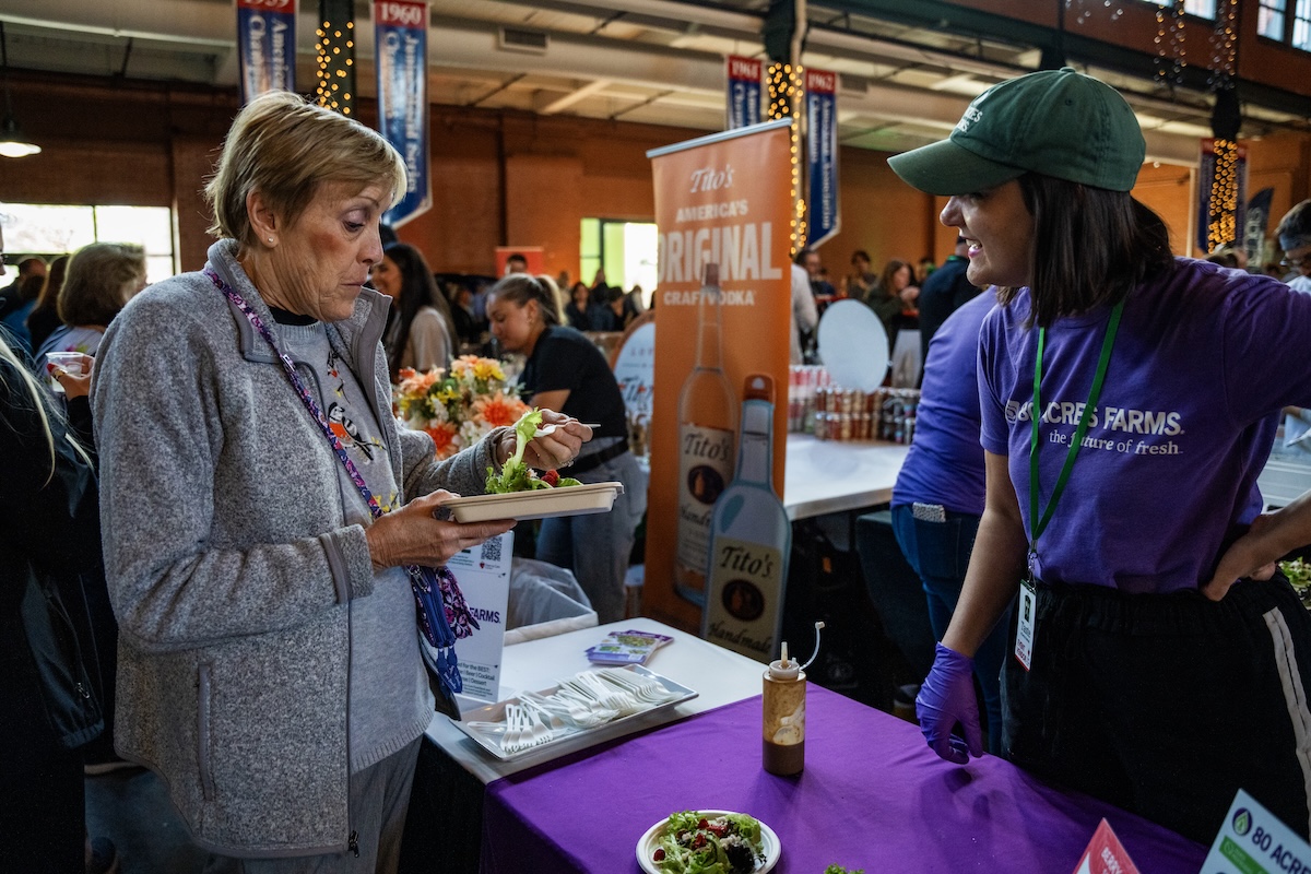 A woman in a grey fleece jacket samples a small salad from a cardboard tray at an indoor food event. She stands across a purple-clothed table from a representative in a green cap and purple "80 Acres Farms" t-shirt, who is smiling and talking to her. In the background, a tall orange banner for "Tito's Handmade Vodka" is visible among a crowd of people in a large, industrial-style hall.