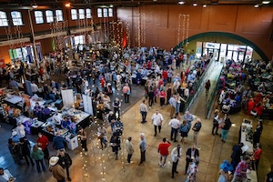 A high-angle, wide shot of a large, crowded indoor event space with high ceilings and industrial-style architecture. Dozens of people are milling around numerous vendor booths and tables spread across the floor. String lights hang from the ceiling, and large windows along the top left wall let in natural light. To the right, a long ramp leads up towards a large arched doorway.