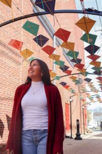 A medium shot of a smiling woman with long dark hair, wearing a white turtleneck sweater and a deep red open cardigan with blue jeans. She is looking upward with a joyful expression while standing in a narrow alleyway with red brick walls. Above her, dozens of colorful diamond-shaped kites in shades of green, yellow, orange, and blue are strung across the alley on black metal arches. The scene is brightly lit with a clear, soft sky visible at the end of the alley.