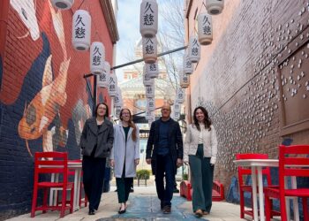 A low-angle, full-length shot of four smiling people—three women and one man—walking toward the camera through Kuji Alley in Franklin, Indiana. They are dressed in business-casual coats and trousers. The narrow alley features a vibrant blue painted ground and red brick walls adorned with murals of koi fish and white blossoms. Dozens of white Japanese lanterns with black kanji characters are strung overhead between the buildings. Red chairs and white tables line the walkway, and a historic brick building with a clock tower is visible at the end of the alley.