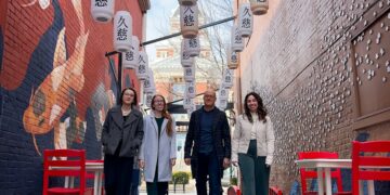 A low-angle, full-length shot of four smiling people—three women and one man—walking toward the camera through Kuji Alley in Franklin, Indiana. They are dressed in business-casual coats and trousers. The narrow alley features a vibrant blue painted ground and red brick walls adorned with murals of koi fish and white blossoms. Dozens of white Japanese lanterns with black kanji characters are strung overhead between the buildings. Red chairs and white tables line the walkway, and a historic brick building with a clock tower is visible at the end of the alley.
