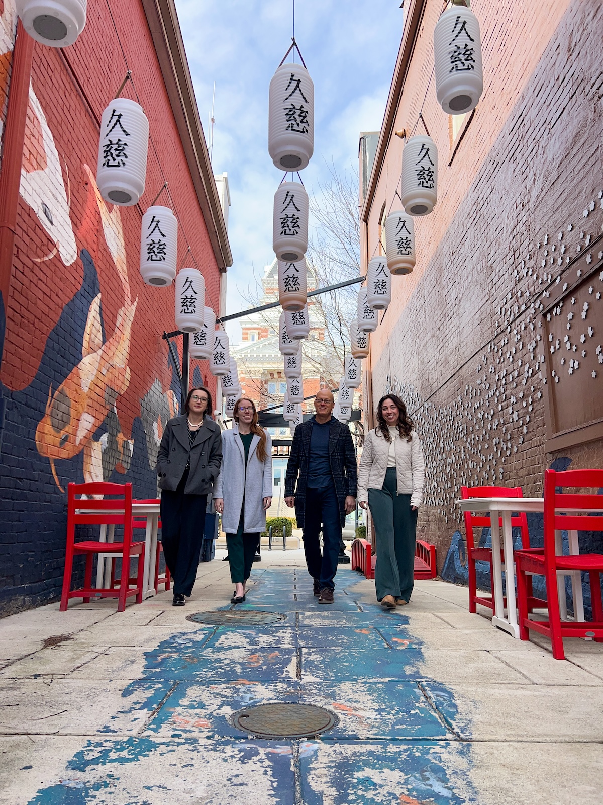 A low-angle, full-length shot of four smiling people—three women and one man—walking toward the camera through Kuji Alley in Franklin, Indiana. They are dressed in business-casual coats and trousers. The narrow alley features a vibrant blue painted ground and red brick walls adorned with murals of koi fish and white blossoms. Dozens of white Japanese lanterns with black kanji characters are strung overhead between the buildings. Red chairs and white tables line the walkway, and a historic brick building with a clock tower is visible at the end of the alley.