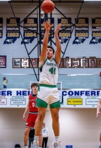 A high-action shot of a high school basketball game. A player from Yorktown, wearing a white jersey with green trim and the number 41, is captured mid-air as he shoots a basketball. Behind him, a player in a red jersey watches the shot. The background features a gymnasium wall adorned with numerous blue and white state championship banners and a conference sign listing several local schools.