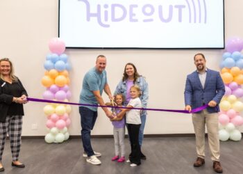 A large group celebrates a ribbon-cutting ceremony in front of a screen displaying a logo that says "HiDEOUT." The Action: In the center, a man, a woman, and two young girls all hold large ceremonial scissors together, poised to cut a long purple ribbon. The Supporters: On the far left, a woman in a black top and plaid pants holds one end of the ribbon, while a man in a blue blazer and tan slacks holds the other end on the right. The Decor: The group is framed by two tall pillars of colorful pastel balloons in shades of pink, blue, orange, and white.