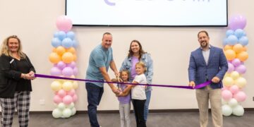 A large group celebrates a ribbon-cutting ceremony in front of a screen displaying a logo that says "HiDEOUT." The Action: In the center, a man, a woman, and two young girls all hold large ceremonial scissors together, poised to cut a long purple ribbon. The Supporters: On the far left, a woman in a black top and plaid pants holds one end of the ribbon, while a man in a blue blazer and tan slacks holds the other end on the right. The Decor: The group is framed by two tall pillars of colorful pastel balloons in shades of pink, blue, orange, and white.