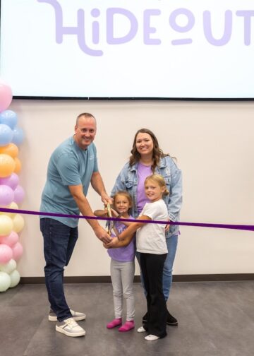 A large group celebrates a ribbon-cutting ceremony in front of a screen displaying a logo that says "HiDEOUT." The Action: In the center, a man, a woman, and two young girls all hold large ceremonial scissors together, poised to cut a long purple ribbon. The Supporters: On the far left, a woman in a black top and plaid pants holds one end of the ribbon, while a man in a blue blazer and tan slacks holds the other end on the right. The Decor: The group is framed by two tall pillars of colorful pastel balloons in shades of pink, blue, orange, and white.
