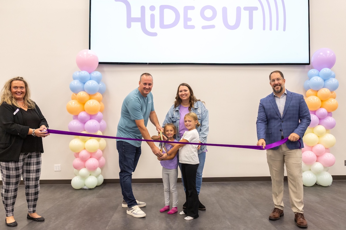 A large group celebrates a ribbon-cutting ceremony in front of a screen displaying a logo that says "HiDEOUT." The Action: In the center, a man, a woman, and two young girls all hold large ceremonial scissors together, poised to cut a long purple ribbon. The Supporters: On the far left, a woman in a black top and plaid pants holds one end of the ribbon, while a man in a blue blazer and tan slacks holds the other end on the right. The Decor: The group is framed by two tall pillars of colorful pastel balloons in shades of pink, blue, orange, and white.