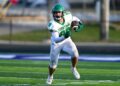 A Yorktown Tigers football player, wearing number 88, runs down a green turf field during a game. He is dressed in a white jersey with green lettering, white pants, a green helmet, and green gloves, firmly clutching the football. The background shows a blurred view of the sidelines under bright daylight.