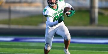 A Yorktown Tigers football player, wearing number 88, runs down a green turf field during a game. He is dressed in a white jersey with green lettering, white pants, a green helmet, and green gloves, firmly clutching the football. The background shows a blurred view of the sidelines under bright daylight.