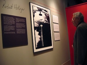 A visitor in a dark suit stands in an art gallery, looking closely at a black-and-white photograph on a neutral-colored wall.
