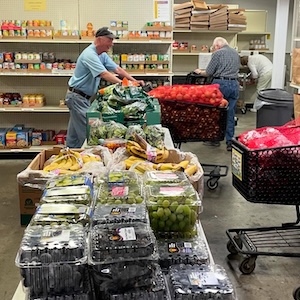This image shows three people working in a busy food pantry or warehouse, surrounded by a large quantity of fresh produce and shelves of canned goods.