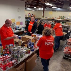 Five volunteers in bright orange "WAVE" shirts and one in black are busy sorting and packing food in a large, industrial pantry. In the foreground, a man at a long table packs cardboard boxes with a variety of canned goods, pasta, and juice bottles. Behind him, other volunteers are gathered around workstations, organizing items against a backdrop of metal shelves stocked with food supplies.