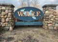 A large, oval-shaped entrance sign for the Wawasee Area Conservancy Foundation (WACF), mounted between two rustic stone pillars. The sign is dark teal with "W.A.C.F." written in bold gold letters over a light blue map of Lake Wawasee. Below the oval, a rectangular plaque displays the full name of the organization. The sign is surrounded by dry, dormant grass and tall reeds under a cloudy sky.