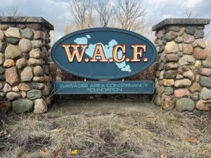 A large, oval-shaped entrance sign for the Wawasee Area Conservancy Foundation (WACF), mounted between two rustic stone pillars. The sign is dark teal with "W.A.C.F." written in bold gold letters over a light blue map of Lake Wawasee. Below the oval, a rectangular plaque displays the full name of the organization. The sign is surrounded by dry, dormant grass and tall reeds under a cloudy sky.